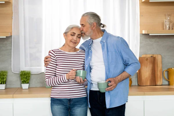 Portrait Of Loving Senior Spouses Relaxing With Coffee In Kitchen Interior, Romantin Elderly Couple Bonding At Home, Caring Man Kissing His Wife To Forehead, Enjoying Spending Time With Each Other