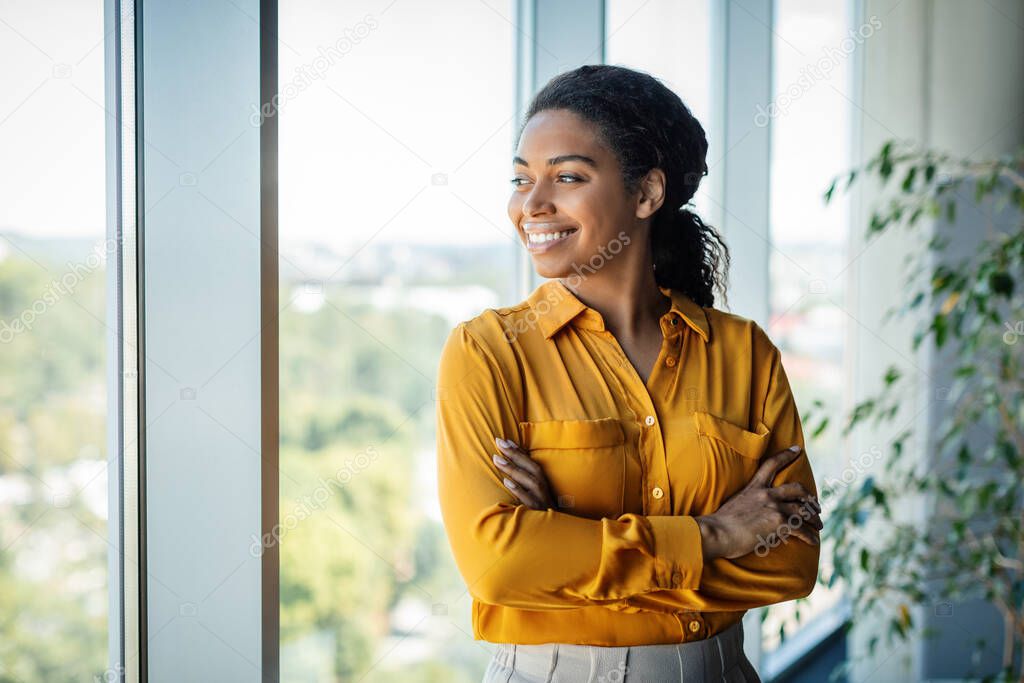 Portrait of confident african american businesswoman standing near window in modern company office, looking away and smiling, free space. Successful female entrepreneur posing with crossed arms