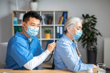Concentrated millennial korean man doctor in protective mask with stethoscope listens to breath of elderly female patient in clinic office interior. Examination, treatment with therapist, health care