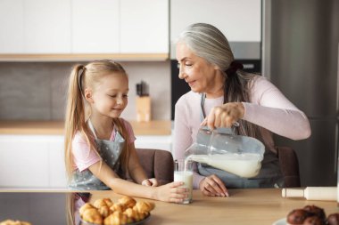 Sweets are ready. Happy european elderly grandmother in apron pours milk at glass of granddaughter at kitchen interior with croissants. Homemade food, cooking together, baking for family at home