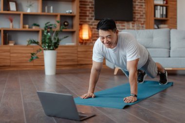 Home workout concept. Mature asian man exercising with laptop computer online standing in plank in living room, free space. Fitness and active lifestyle concept