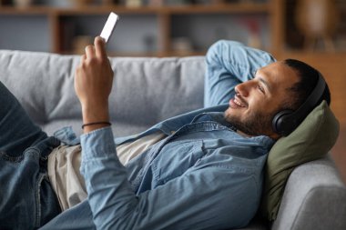 Favorite Leisure. Happy Black Guy In Wireless Headphones Listening Music On Smartphone While Relaxing On Couch At Home, Smiling African American Man Resting On Sofa And Enjoying Favorite Playlist