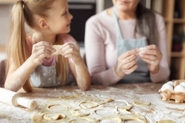 Glad european little granddaughter and elderly grandmother in aprons make cookies from dough in kitchen interior, cropped, close up. Lesson of homemade food at home, chef teaching girl to prepare meal