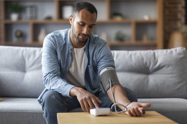 Young Black Man Sitting On Couch And Checking Blood Pressure With Upper Arm Monitor, Millennial African American Guy Measuring Arterial Tension While Relaxing In Living Room At Home, Copy Space