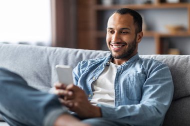 Mobile Communication. Handsome Black Man Messaging On Smartphone While Relaxing On Couch At Home, Smiling Young African American Guy Texting With Friends Or Browsing Social Media, Closeup Shot