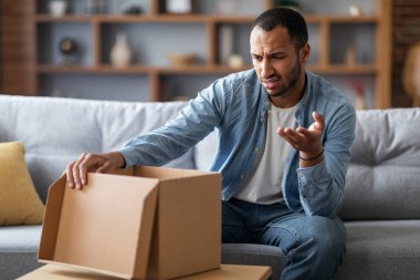 Wrong Parcel. Confused black man looking at open cardboard box at home and frowning, young african american guy disappointed about shipping mistake, feeling upset after bad delivery service