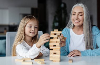 Happy european little girl and old woman play in tower game on table in living room interior. Grandmother and granddaughter relationship, board game and fun together at home during covid-19 quarantine