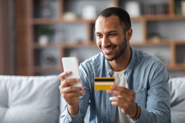 Online Shopping. Handsome Young Black Man Using Smartphone And Credit Card While Resting On Couch At Home, Smiling African American Guy Making Internet Purchases Or Mobile Payments, Copy Space
