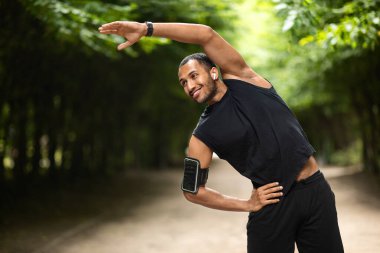 Happy athletic young african american man in black sportswear exercising outdoors, black guy stretching in public park, having workout,listening to music, using erabuds, looking at copy space, smiling