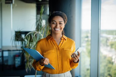 Happy successful businesswoman holding smartphone and clipboard with documents, standing in modern office interior, smiling at camera. Business at workplace