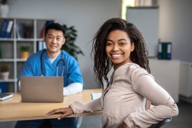 Smiling adult asian male doctor with laptop consultation young african american woman patient, look at camera in clinic office interior. Visit to therapist, treatment of illness, medical health care
