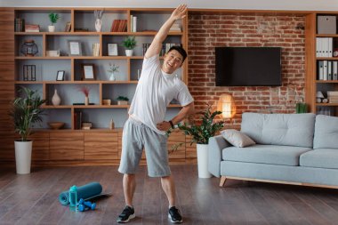 Active mature man warming up and stretching before domestic workout, doing flexibility exercises for healthier living. Man training in living room interior, free space