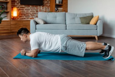 Domestic sports concept. Asian man doing plank exercises at home, training in living room on sports mat, enjoying healthy lifestyle, side view with copy space