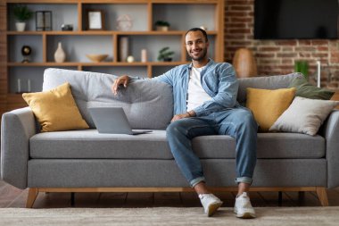 Handsome Young Black Man Sitting On Couch With Laptop And Smiling At Camera, Happy Millennial African American Guy Relaxing With Computer In Cozy Living Room Interior At Home, Copy Space