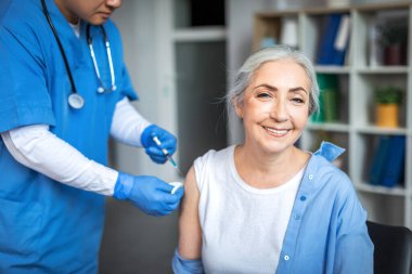 Millennial man doctor make injection to smiling elderly lady patient in clinic office interior. Treatment, medical health care, vaccination and immunization of retired during flu and covid-19 outbreak