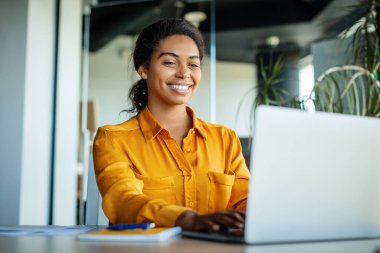 Happy black businesswoman using laptop typing and working online while sitting at workplace in office, copy space. Female manager chatting with clients on computer
