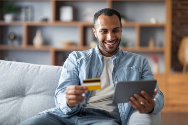 Online Payments. Handsome Black Young Man Using Digital Tablet And Credit Card While Resting On Couch At Home, Cheerful African American Guy Shopping In Internet, Enjoying E-Commerce, Copy Space
