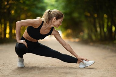 Active blonde middle aged woman stretching after jogging at public park, sporty attractive lady having workout outdoors, enjoying morning activity, panorama with copy space. Open air fitness concept