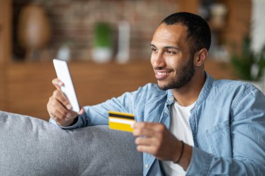 Mobile Payments Concept. Smiling Black Guy Using Smartphone And Credit Card While Sitting On Couch In Living Room At Home, Handsome African American Man Transfer Money Online , Enjoying E-Commerce