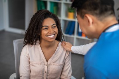 Mature asian man consulting cheerful millennial african american woman patient in clinic office interior. Recovery, check-up with doctor, treatment of illness, health care and great medical service