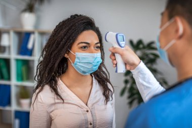 Millennial asian man checks temperature with thermometer of young black lady patient in protective mask in clinic interior. Examination with doctor, treatment, health care during covid-19 pandemic