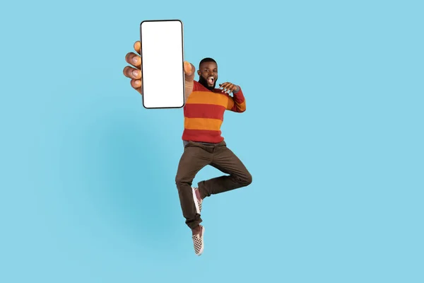 Check This. Excited Black Man Jumping And Showing Big Blank Smartphone, Demonstrating Empty Mobile Phone In Hand, Recommending New Application While Having Fun Over Blue Background, Collage, Mockup