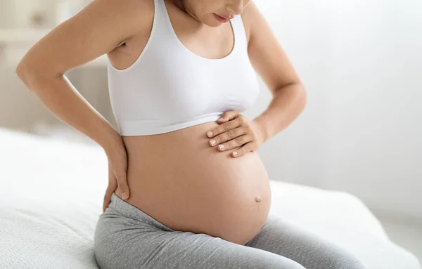 Cropped of expecting woman in pajamas having back pain, sitting on bed at home in the morning, touching ache zone and big tummy, unrecognizable pregnant lady suffering from backache, copy space