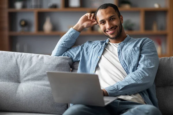 Portrait Of Handsome African American Male Using Laptop While Sitting On Couch At Home, Black Millennial Man Working Remotely On Computer In Living Room And Smiling At Camera, Copy Space