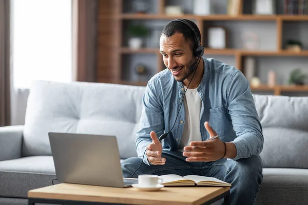 Distance Communication. Smiling Black Guy In Headset Having Web Conference On Laptop At Home, Young African American Man Talking And At Computer Camera While Sitting On Couch In Living Room