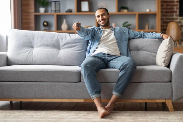 Home Relax. Handsome Black Man Sitting On Couch And Switching Tv Channels, Smiling Young African American Guy Holding Remote Controller, Relaxing On Comfortable Sofa In Living Room, Copy Space