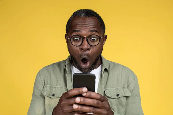 Shocked happy adult african american male in casual with open mouth in glasses looks at smartphone, isolated on yellow background, studio. Ad and offer, emotions, surprise and win, online victory