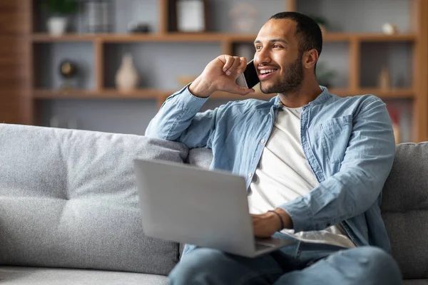 Remote Business. Handsome Black Man Talking On Phone And Using Laptop While Sitting On Couch At Home, African American Freelancer Guy Working On Computer And Enjoying Cellphone Conversation