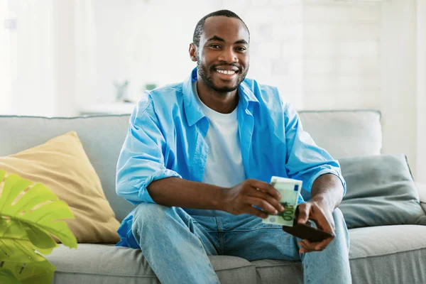 Positive african american guy holding wallet and taking out money, sitting at home on sofa and smiling at camera. Wealthy black man showing cash