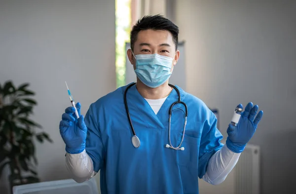 Glad adult japanese man doctor in protective mask and gloves shows syringe and vaccine in clinic interior. Sickness treatment, medical health care, vaccination and immunization due covid-19 pandemic