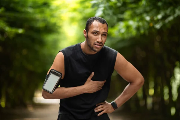 Handsome young african american sportsman having heart pain, touching his chest, jogging in the public park, using armband with smartphone, mockup, copy space. Sport and cardiovascular diseases