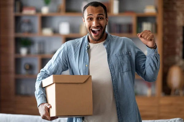 Delivery Concept. Excited Black Man Holding Big Cardboard Box At Home, Joyful Millennial African American Guy Carrying Parcel And Raising Fist, Emotionally Reacting To Fast Shipping, Copy Space