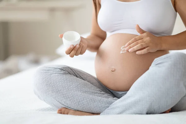 Cropped of pregnant woman holding beauty product, applying moisturizing cream on her belly, sitting on bed at home, panorama with empty space. Stretch mark treatment and cosmetics during pregnancy