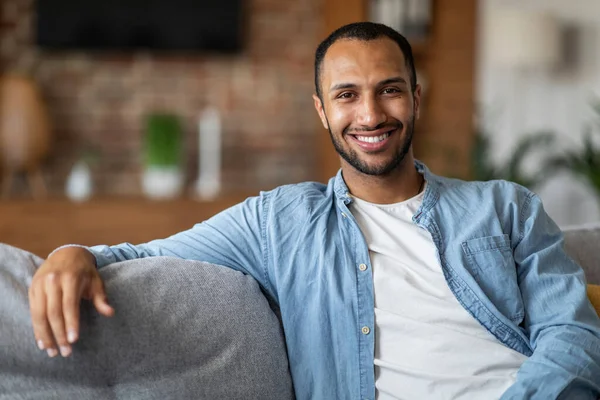 Portrait Of Smiling Young Black Man Posing On Couch At Home, Handsome Millennial African American Guy Resting On Comfortable Sofa In Living Room And Looking At Camera, Enjoying Domestic Leisure