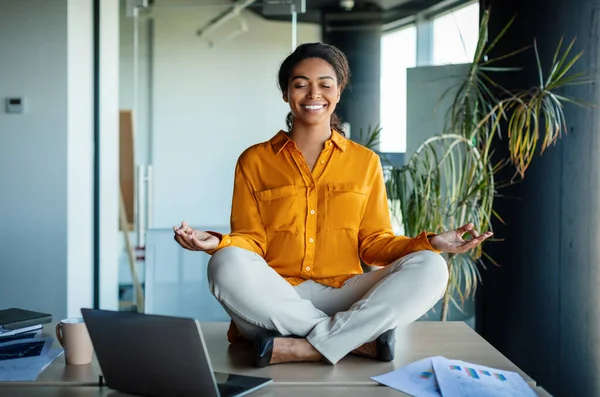 Office zen. Calm black businesswoman meditating with closed eyes on office desk, sitting at workplace in lotus position. Positive woman feeling peaceful and balanced