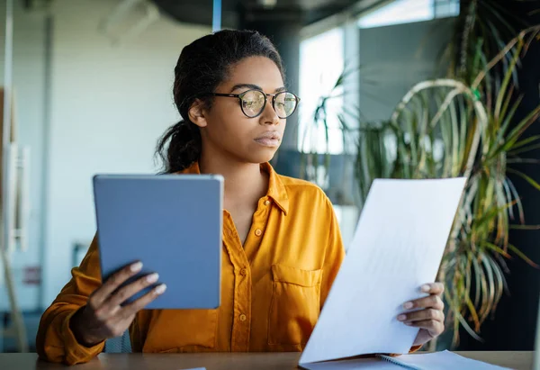 Serious black woman entrepreneur using digital tablet and reading documents while planning companys budget, working through financial papers in her office