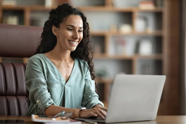 Positive young woman with curly long hair freelancer using laptop at home, lady sitting at workdesk at office, typing on keyboard and smiling, sending emails, copy space. Remote job concept