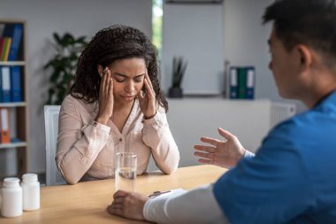 Serious adult asian male doctor calms sad young african american female patient in clinic office interior. Client support, examination with therapist, treatment of illness and medical health care