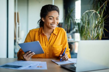 Positive black woman using digital tablet and laptop computer, working online in office, having video call with colleagues. Female entrepreneurship, gadgets and technology concept