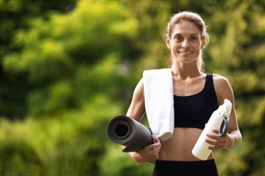 Cheerful athletic middle aged woman in black sportswear having yoga at public park, holding towel, fitness mat, bottle of water, having break while training, panorama with copy space. Fitness outdoos