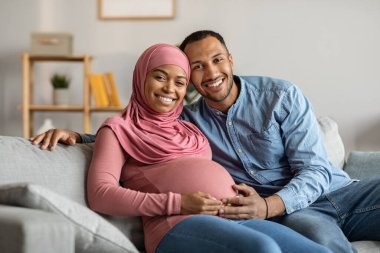 Portrait Of Happy Black Muslim Couple Expecting Baby Posing In Home Interior, Joyful Young Islamic Spouses Sitting On Couch In Living Room And Smiling At Camera, Husband Touching Wifes Belly
