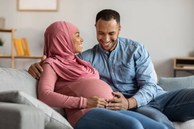 Portrait Of Happy Islamic Couple Expecting Baby Relaxing Together At Home, Pregnant Black Muslim Spouses Embracing On Couch, Smiling Husband And Wife Resting In Living Room, Free Space