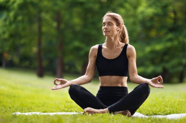 Peaceful blonde woman in black tight sportswear sitting on fitness mat with closed eyes in lotus pose, practicing yoga at public park, listening to music, using brand new earbuds, copy space