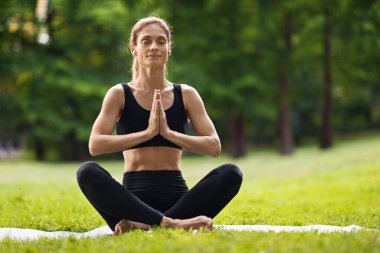 Calm athletic good-looking blonde middle aged woman meditating in public park, yoga lady sitting on fitness mat in green forest, listening to music, using earbuds, panorama with copy space