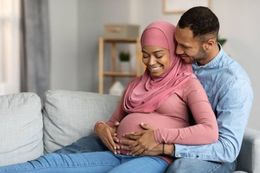 Loving Husband Hugging Pregnant Muslim Wife While They Relaxing On Couch At Home, Black Islamic Spouses Expecting Baby Resting Together In Living Room Interior, Enjoying Upcoming Parenthood