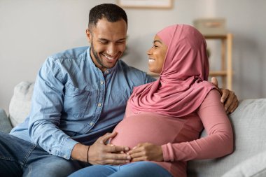 Happy Expecting Muslim Couple Relaxing Together On Sofa In Living Room, Loving Husband Tenderly Touching Wifes Belly, Cheerful Pregnant Islamic Spouses Bonding At Home, Closeup Portrait